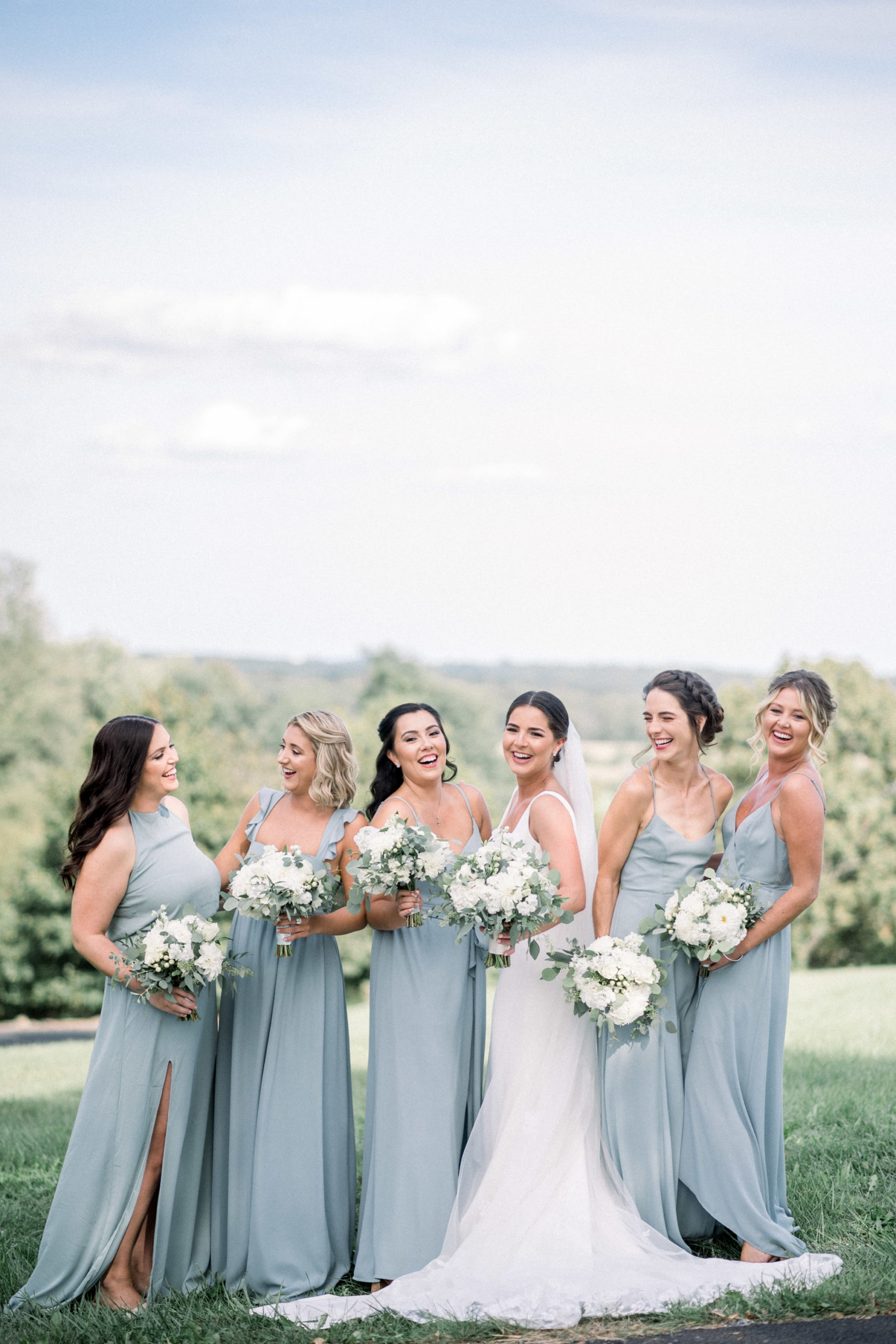 bride standing with her bridesmaids on a hilltop as they all laugh together