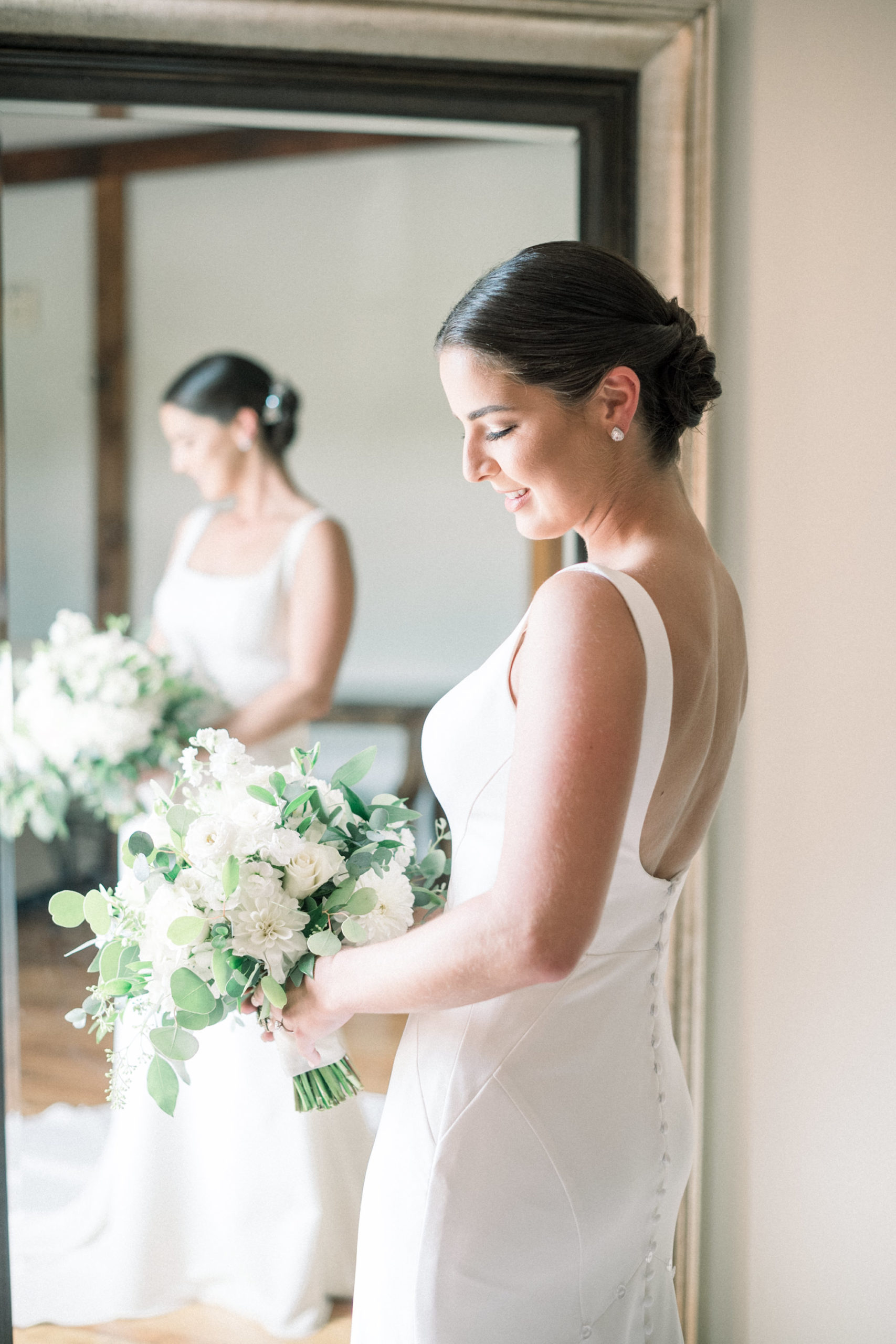 bride with a low bun and a satin wedding dress holding a white floral wedding bouquet before her wedding ceremony 