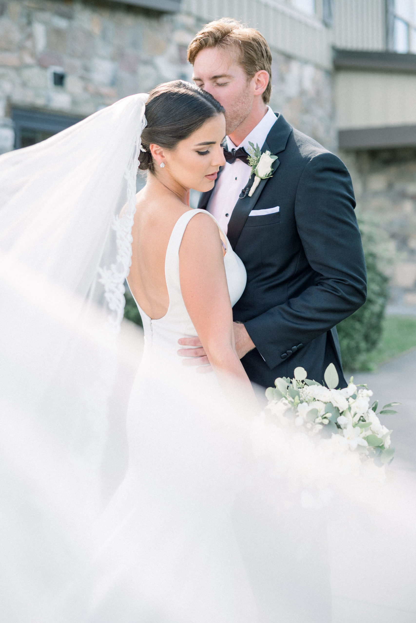 groom holds his bride as her veil blows in the wind and she looks down her shoulder