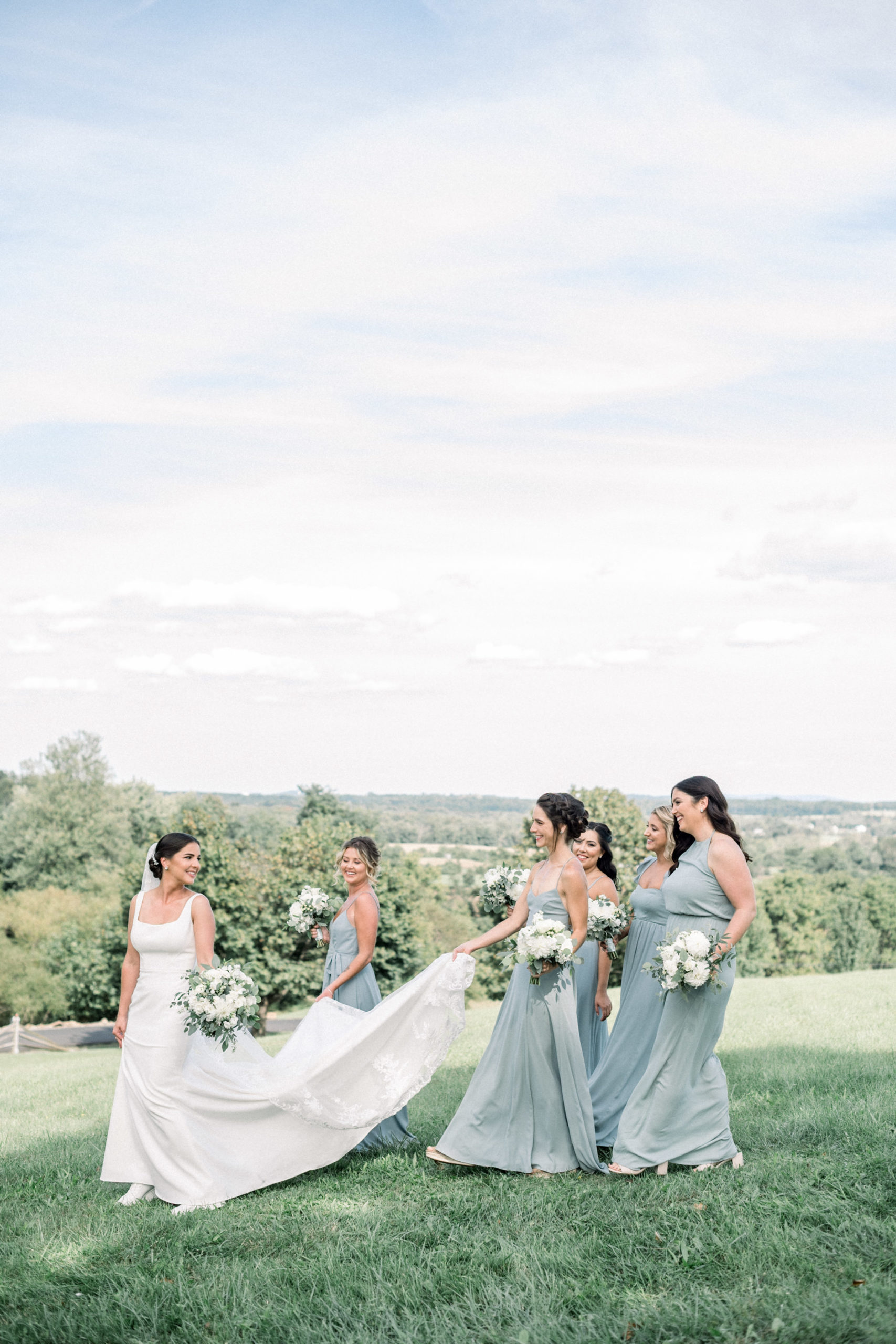 bride walking down a hill while her bridesmaids carry her train down the hill for her outdoor summer wedding